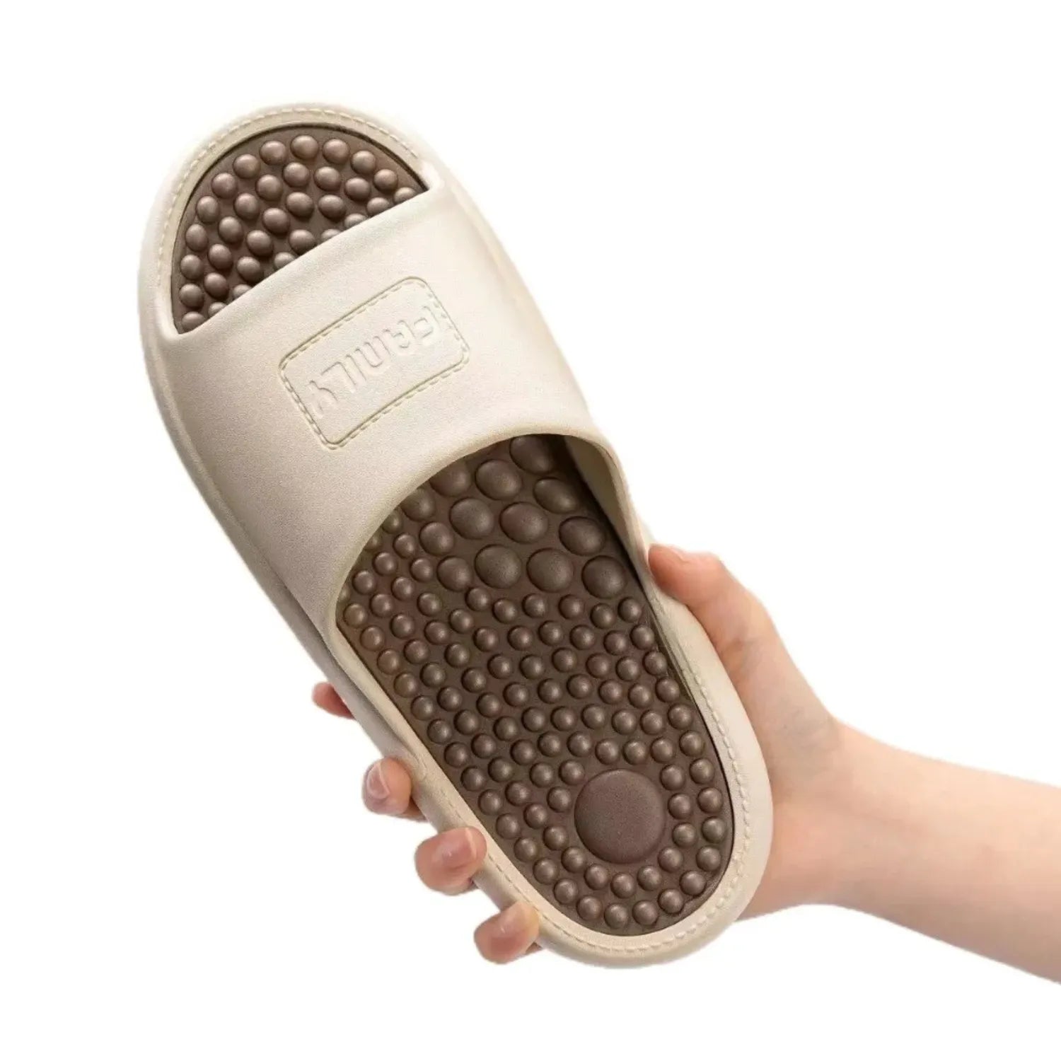 Uma mão segura um chinelo de massagem na cor marrom. O calçado tem uma sola com pontos de massagem elevados e um design antiderrapante para conforto e terapia nos pés.